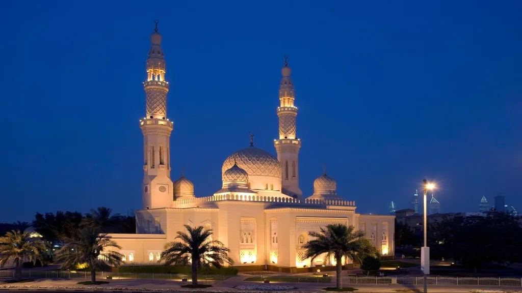 Large mosque with glowing golden lights, two tall minarets, domes, and palm trees against a deep blue evening sky.