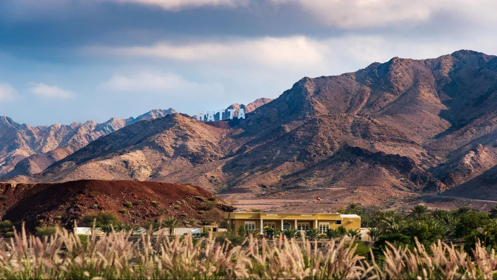 Rocky mountains with large white “HATTA” letters on the slope, a yellow building with a flag at the base, and greenery in the foreground.