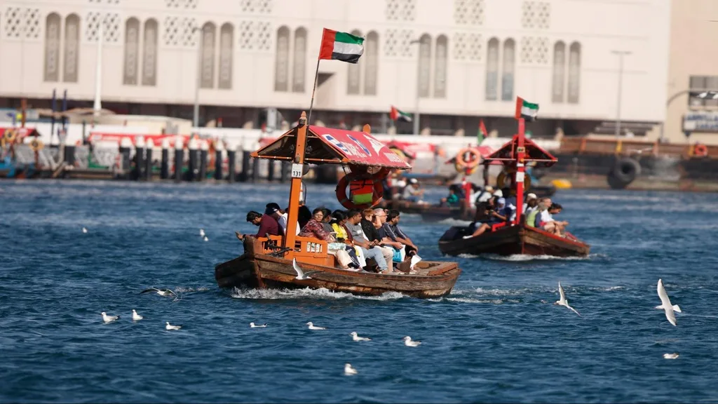 Wooden passenger boats with UAE flags crossing Dubai Creek, surrounded by seagulls and a dockside building in the background.
