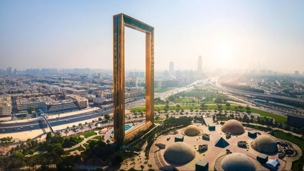 Massive golden rectangular Dubai Frame structure in Zabeel Park, with landscaped gardens and city skyscrapers in the hazy background.