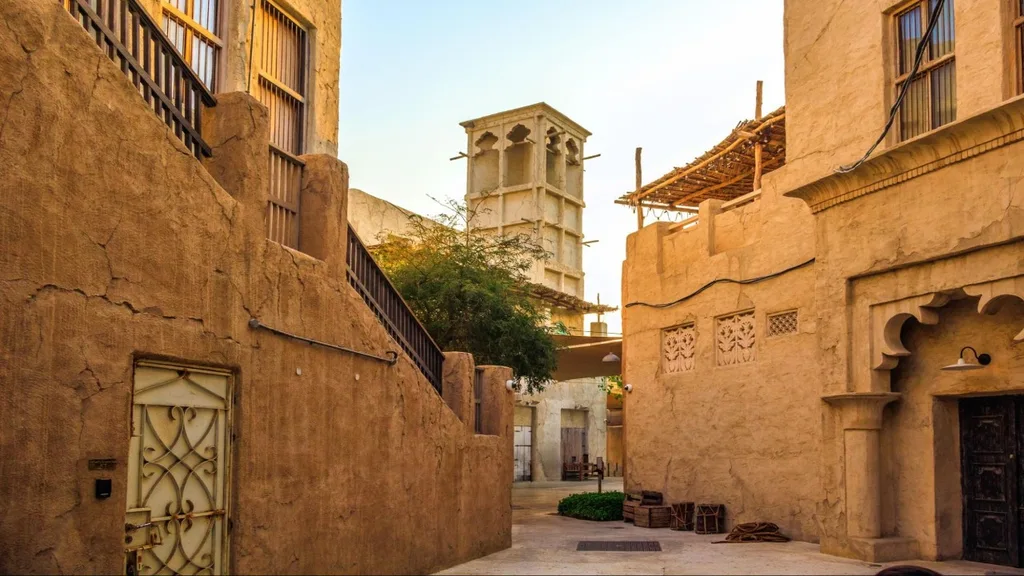 Narrow alleyway with sand-colored buildings, wooden doors, and a tall wind tower in the background, lit by warm evening sunlight.